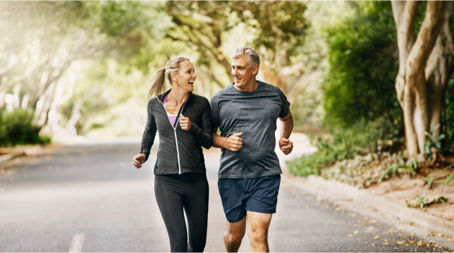 A man and woman jogging on a road in the early morning, surrounded by trees and a clear blue sky.